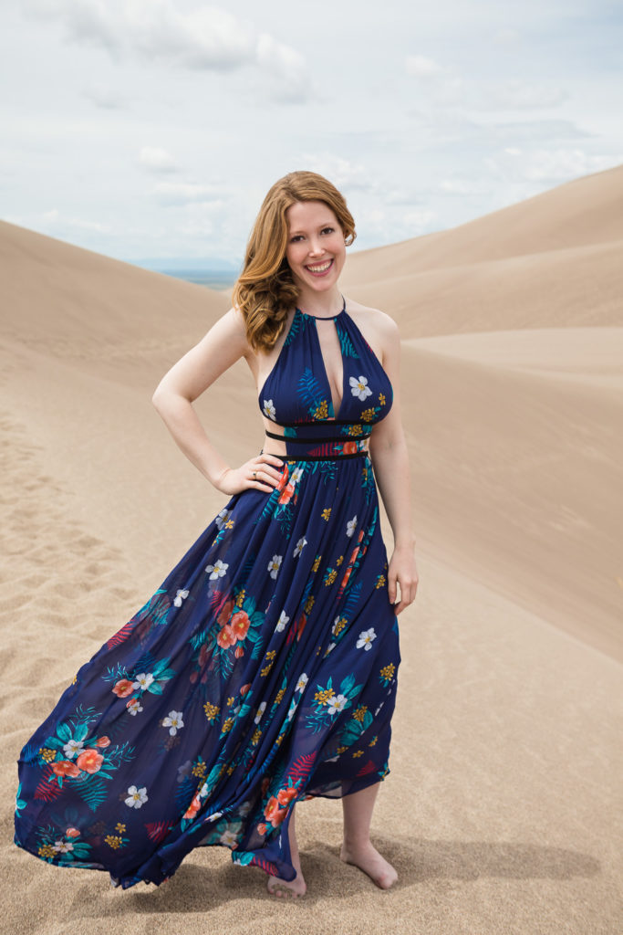Melissa is wearing a long blue dress with a tropical floral pattern and cut-outs at her waist. One hand is resting on her hip while the other is relaxed by her side. She is standing on sand dunes and the sky is overcast with layered clouds.